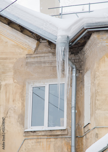 Dangerous, frozen icicles hang on a drainpipe, spring or winter. Wall of an old brick house with windows. Large cascades of icicles in smooth, beautiful rows. Cloudy winter day, soft light.