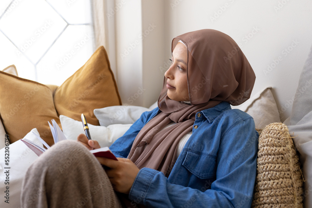 Happy biracial woman wearing hijab, sitting on sofa in living room, taking notes