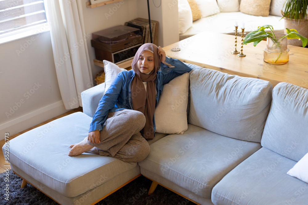 Happy biracial woman wearing hijab, sitting on sofa in living room Happy biracial woman wearing hijab, sitting on sofa in living room