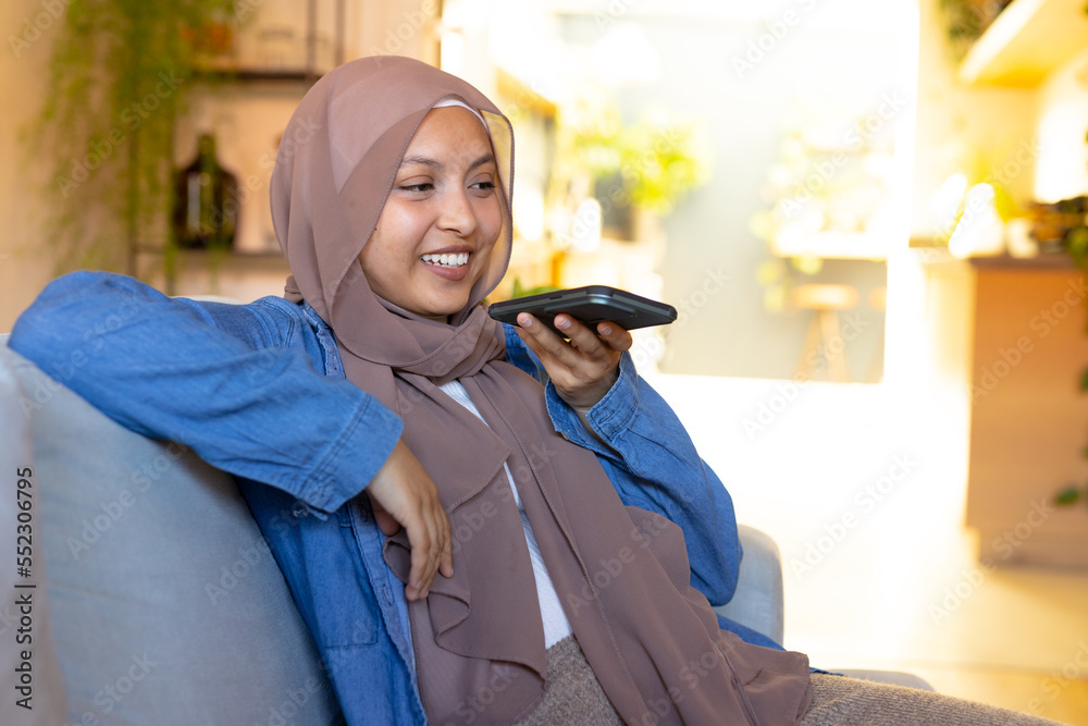 Happy biracial woman wearing hijab, sitting on sofa in living room, talking on smartphone Happy biracial woman wearing hijab, sitting on sofa in living room, talking on smartphone