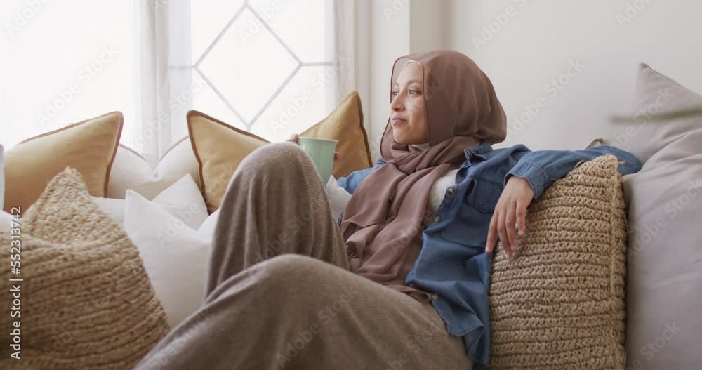 Video of smiling biracial woman in hijab drinking coffee relaxing on sofa at home Video of smiling biracial woman in hijab drinking coffee relaxing on sofa at home