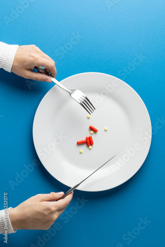 Vertical of hands holding fork and knife over plate with pills, on blue background with copy space