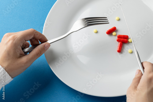 Composition of hands holding fork and knife over plate with pills, on blue background
