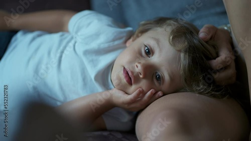 Closeup child face laying on parent leg. Mother caressing small blond boy hair. Mom and kid affection