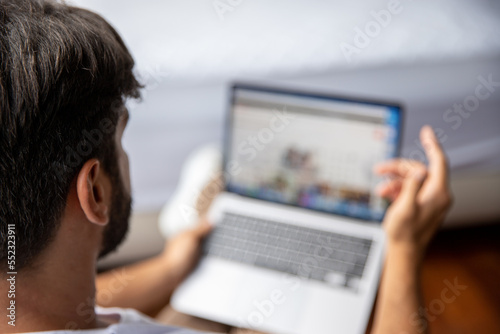 Young tech user relaxing on sofa in front of laptop with screen being shown out of focus. Man surfing the internet, reading news, distance online study work concept. Over-the-shoulder view.