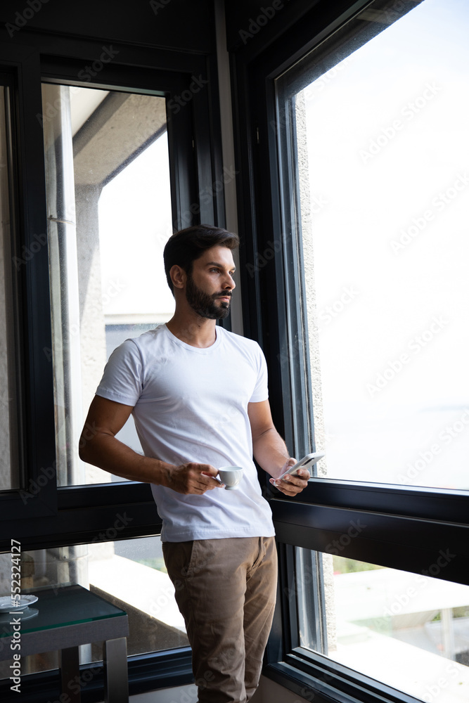 man holding a cup of coffee standing by a window holding his phone looking out the window man holding a cup of coffee standing by a window holding his phone looking out the window