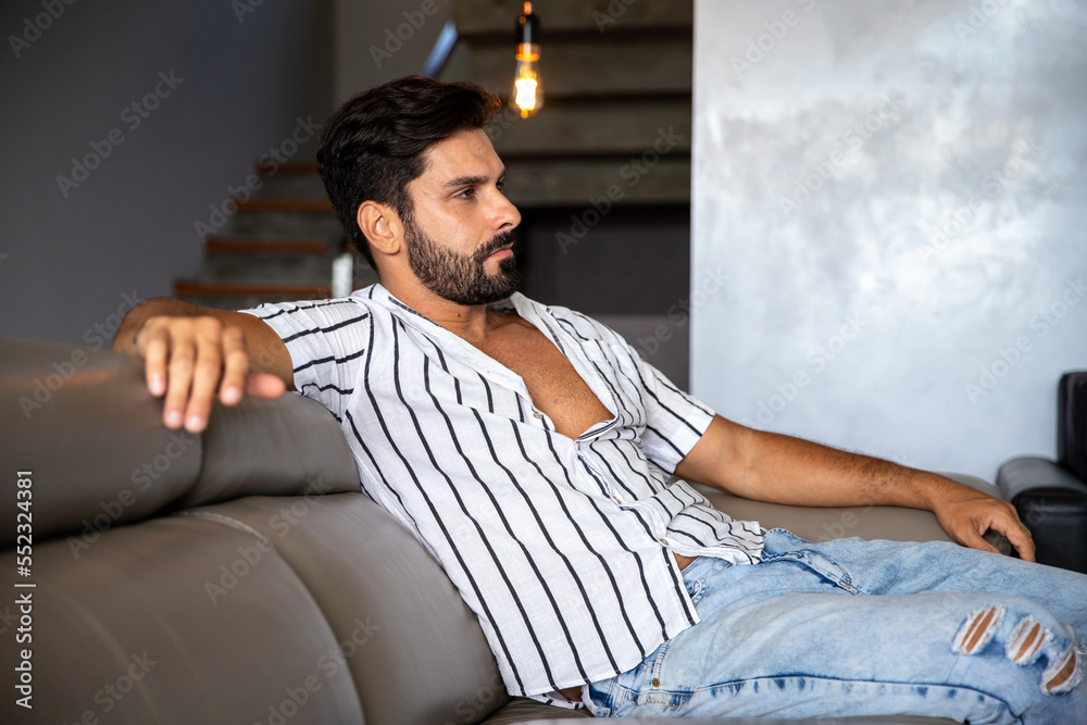 young boy with beard sitting on sofa at home pensive, man at home worried, looking ahead