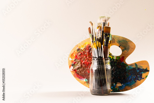 Composition of jar of painting brushes and palette on white background with copy space