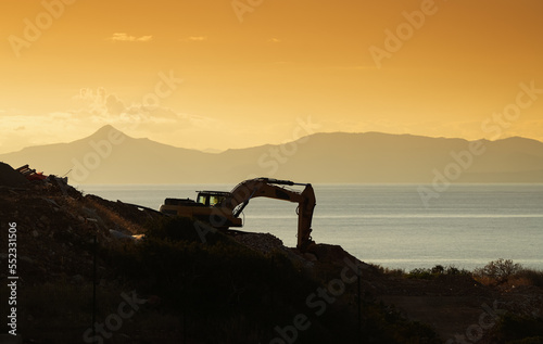 Silhouette of an excavator digging on the sea shore against sunset sky. Construction industry heavy machines.