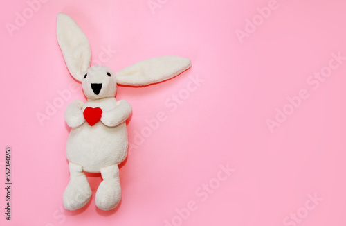 children's toy soft fluffy white rabbit holding a red heart on a pink background