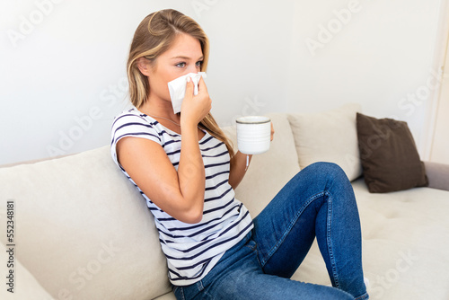Sick young woman blowing running nose, feeling unhealthy and ill, upset female holding paper napkin, handkerchief, holding tea or coffee mug, sitting on couch at home.