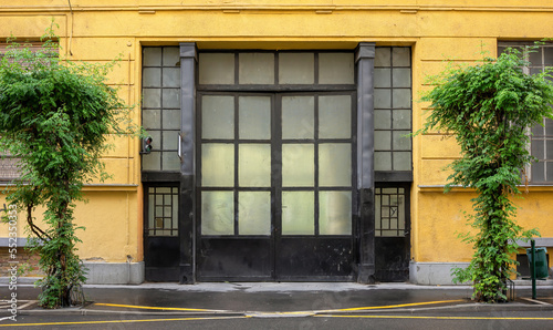 Frontal view of a yellow factory facade with a nostalgic black gate with glass panes in Budapest Hungary. In the foreground is a wet road and two street lamps overgrown with ivy photo