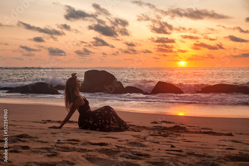Lonely young woman sitting on sandy beach at evening sea waves background, relaxing looking away. Pretty lady wear black dress on tropical ocean at sunset. Travel vacation concept. Copy text space © Alex Vog