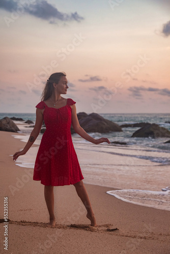 Pretty young woman standing on sandy beach at evening sea background at sunset  looking away. Cute slim lady wear red dress on tropical dusk ocean at sunrise. Travel vacation concept. Copy text space