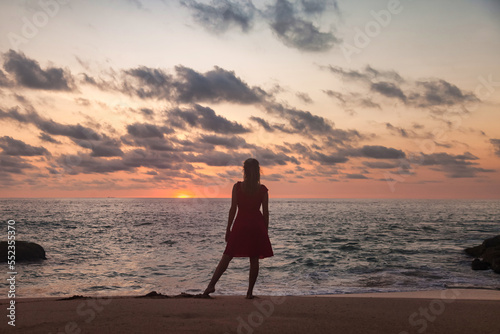 Rear view silhouette young woman standing on sandy beach at evening sea background at sunset. From behind slim lady wear red dress on tropical dusk ocean. Travel vacation concept. Copy text space