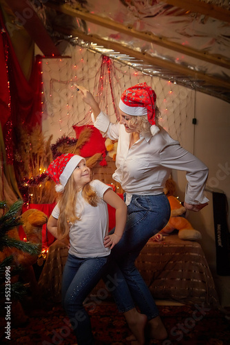 Cute mother and daughter in the red hats of Santa Claus assistants in a room decorated for Christmas. The tradition of decorating house and dressing up for the holidays. Happy childhood and motherhood