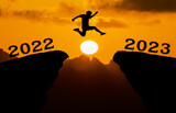 A young man jump between 2022 and 2023 years over the sun and through on the gap of hill silhouette evening colorful sky. happy new year 2023.	