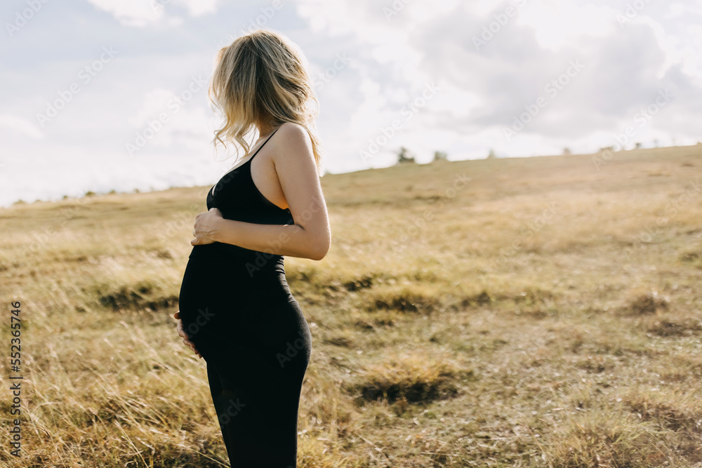Blonde pregnant woman wearing all black standing in an open field, holding hands on belly. Blonde pregnant woman wearing all black standing in an open field, holding hands on belly.