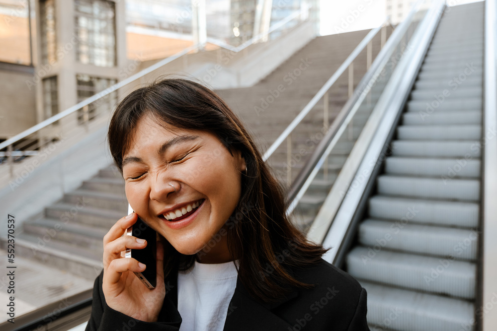 Cheerful asian girl talking on smartphone while standing on escalator outdoors Cheerful asian girl talking on smartphone while standing on escalator outdoors