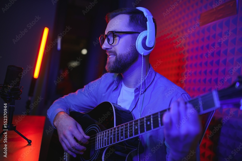 Obraz premium A young stylish guy with a beard wearing casual or music teacher playing guitar in front of smartphone camera. Online guitar training or vocal lesson