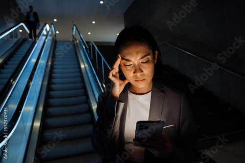 Canvas Print Young asian woman frowning while using smartphone standing on escalator indoors