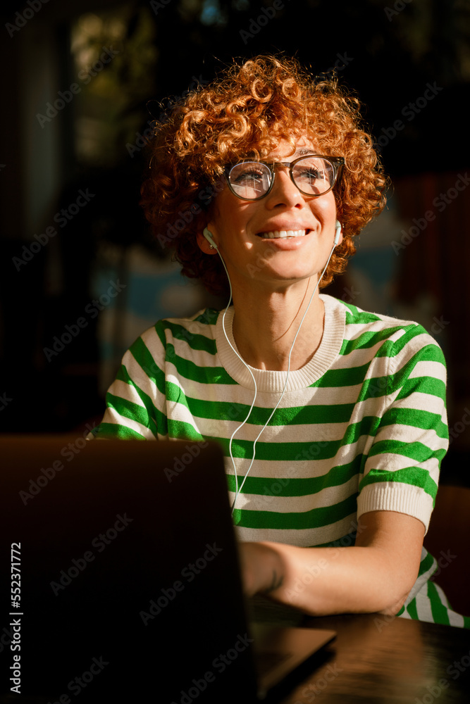 Smiling middle-aged woman in earphones and using laptop while sitting in cafe Smiling middle-aged woman in earphones and using laptop while sitting in cafe