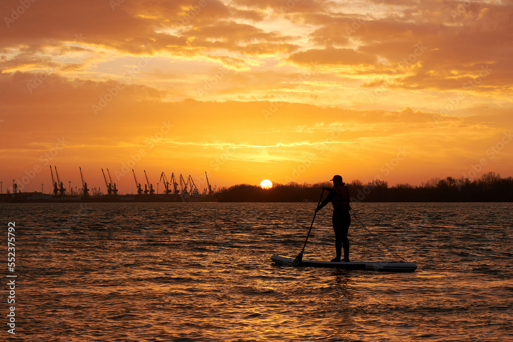 Silhouette of a woman standing on an inflatable SUP board at sunrise and paddling through the shiny water surface of river
