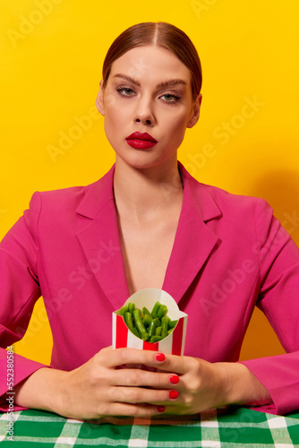 Emotionless, serious young woman in bright pink jacket holding green beans over yellow background. Healthy diet. Food pop art photography.