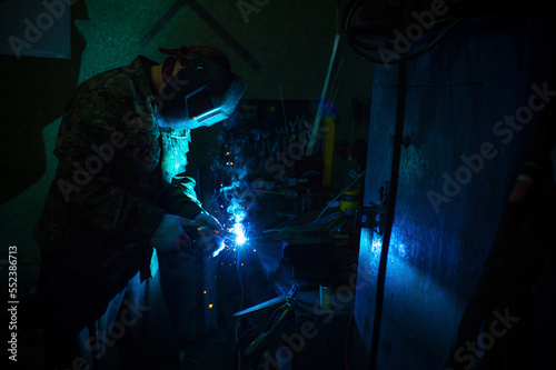 A masked worker performs spot welding work in his workshop, clamping a workpiece in a vise on a workbench © Алексей Вяткин