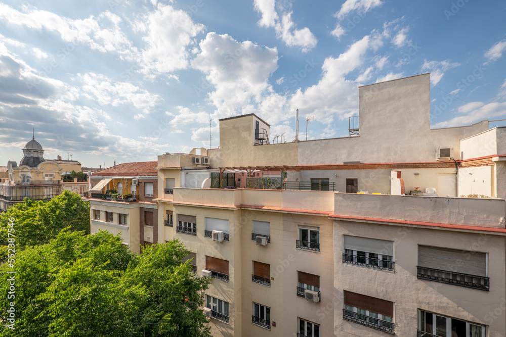 Facades of buildings in a street with leafy trees on a day with some clouds in the sky Facades of buildings in a street with leafy trees on a day with some clouds in the sky