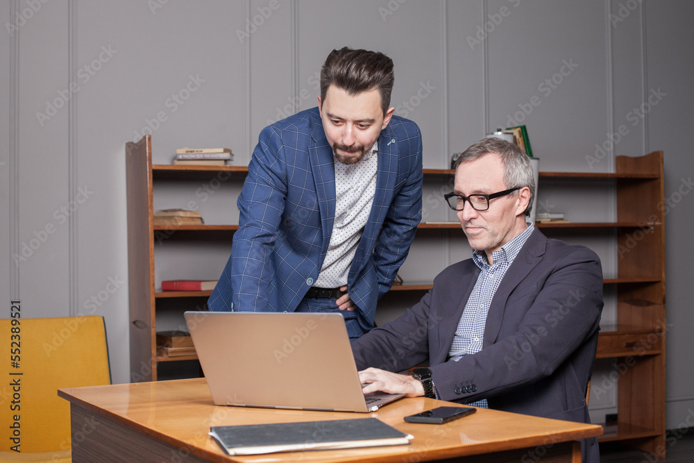 Mature businessman in blue suit and his younger colleague are working on the laptop in the business office Mature businessman in blue suit and his younger colleague are working on the laptop in the business office