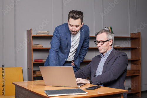 Mature businessman in blue suit and his younger colleague are working on the laptop in the business office