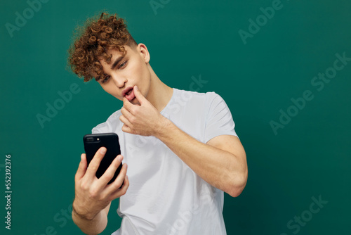 a funny man with curly hair stands on a green background in a gray T-shirt and holds his hand near his mouth, holding the phone in his hand looking into it