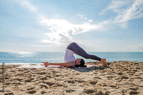 Caucasian flexible woman practicing yoga in plow pose, halasana exercise, yin yoga snail posture at the beach. Side view, working out on the sand outdoors. Lady wearing sportswear doing Plough pose.  photo