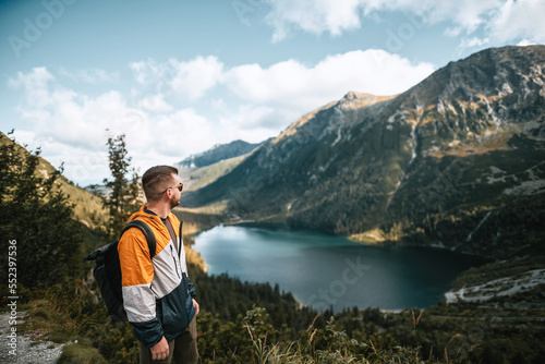 Bearded young caucasian man on the mountain top. Background mountain landscape and lake at vibrant sunset. A man standing on a stone cliff over the lake. Concept of success, freedom and strength.