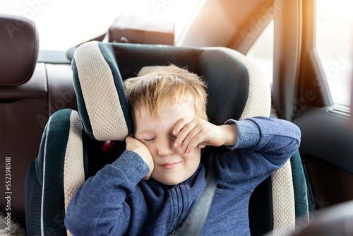 Cute caucasian toddler boy awaken and rub eyes in child safety seat in car during road trip. Adorable baby dreaming asleep comfortable chair during journey in vehicle. Children care safety on roads photo