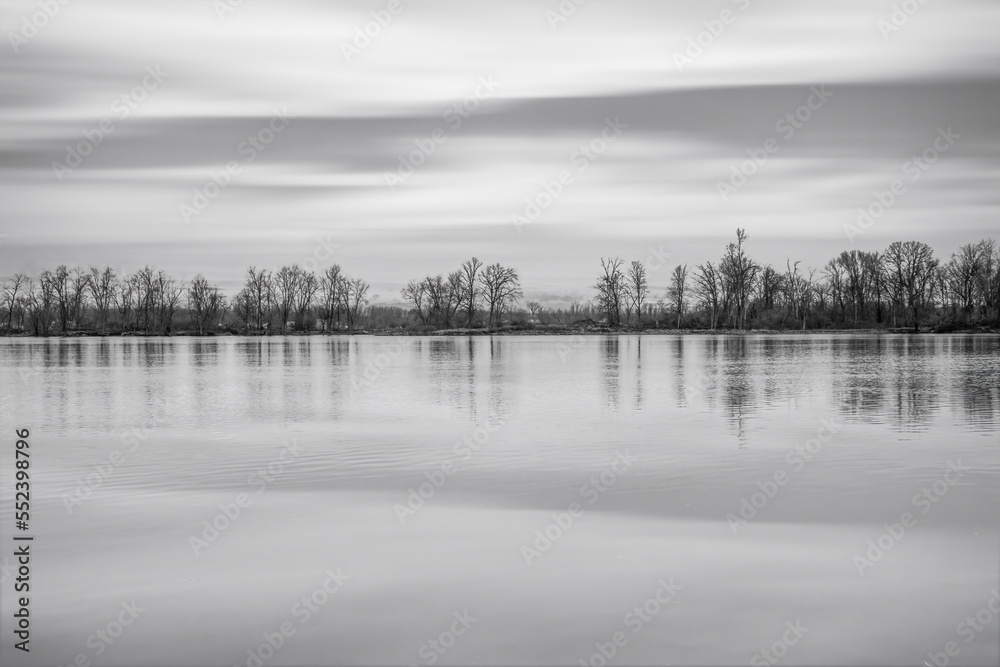 Fototapeta premium View across a calm river at a treed island, sky and trees reflected in water, nobody