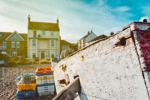 Shallow focus of an old wooden fishing boat showing the flaky paintwork, seen on a shingle beach. Town houses overlooking the beach are in the background.