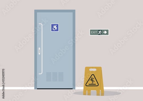 A toilet for wheelchair users, a yellow caution wet floor sign, and a green sticker pointing the evacuation route