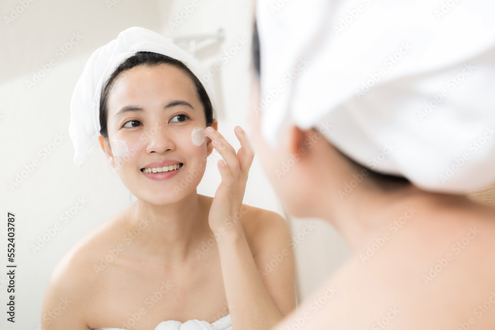 Happy young Asian woman applying face lotions while wearing a towel and touching her face in bathroom Happy young Asian woman applying face lotions while wearing a towel and touching her face in bathroom