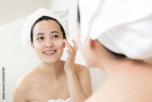 Happy young Asian woman applying face lotions while wearing a towel and touching her face in bathroom