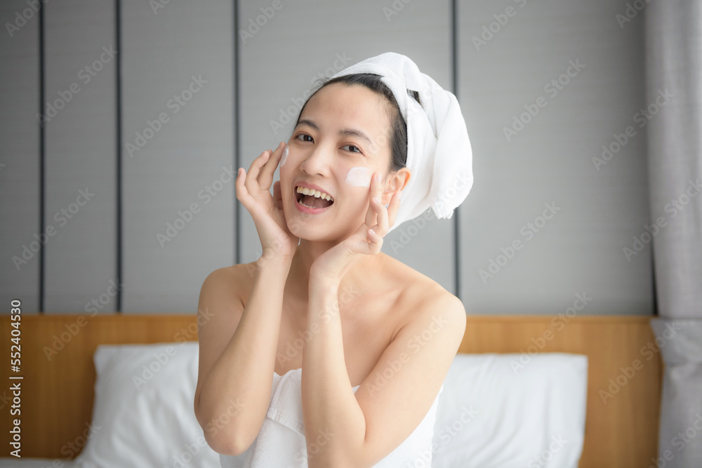 Happy young Asian woman applying face lotions while wearing a towel and touching her face. Daily makeup and skincare Happy young Asian woman applying face lotions while wearing a towel and touching her face. Daily makeup and skincare