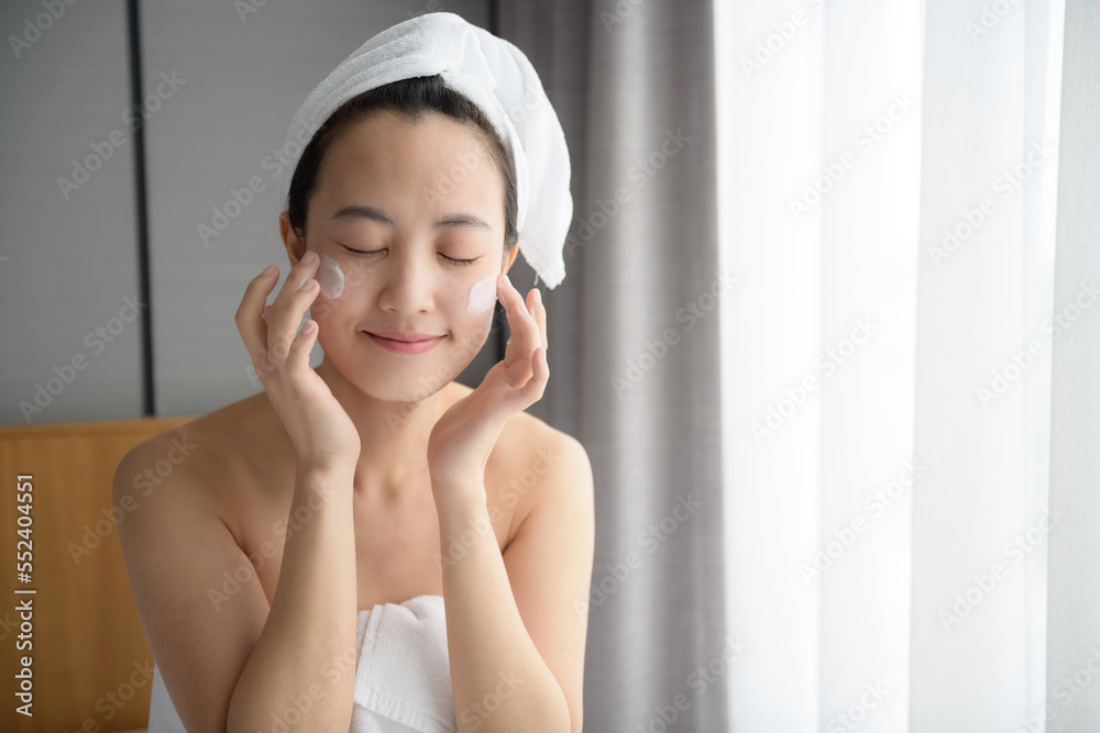 Happy young Asian woman applying face lotions while wearing a towel and touching her face. Daily makeup and skincare Happy young Asian woman applying face lotions while wearing a towel and touching her face. Daily makeup and skincare