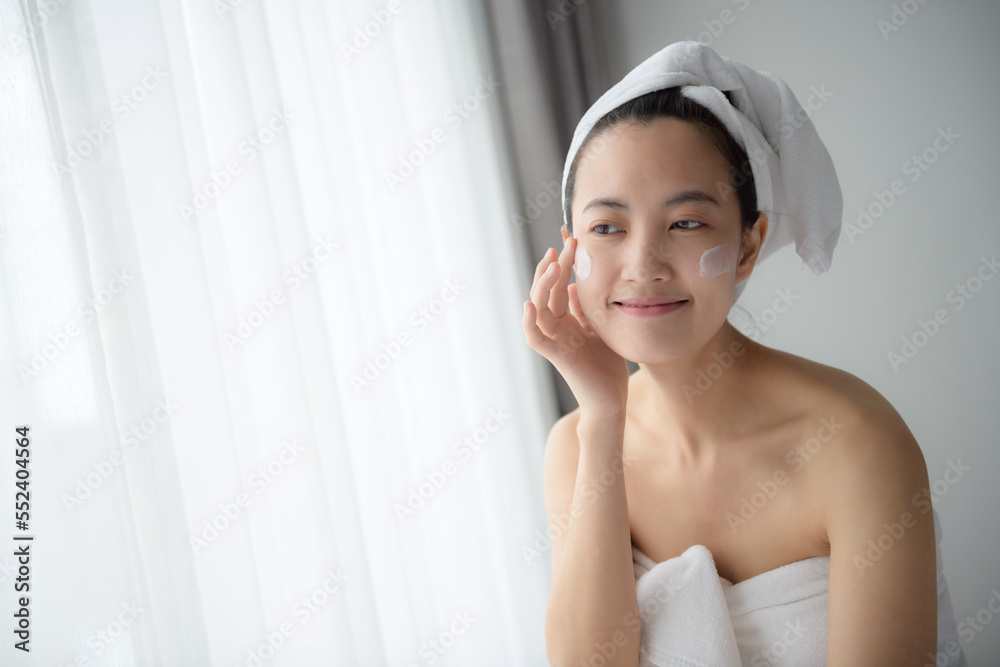 Happy young Asian woman applying face lotions while wearing a towel and touching her face. Daily makeup and skincare Happy young Asian woman applying face lotions while wearing a towel and touching her face. Daily makeup and skincare
