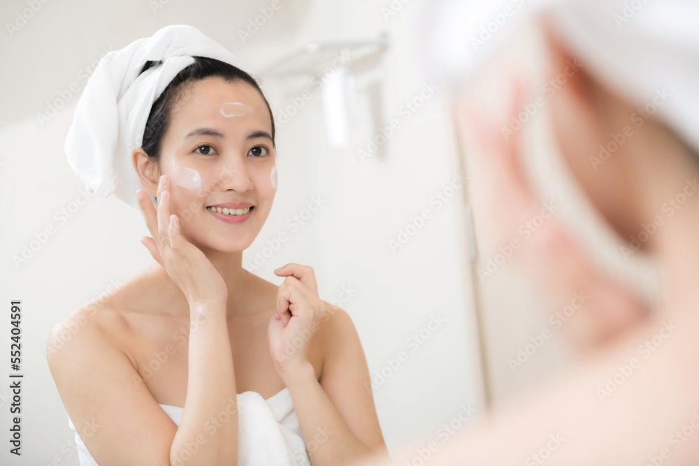 Happy young Asian woman applying face lotions while wearing a towel and touching her face in bathroom Happy young Asian woman applying face lotions while wearing a towel and touching her face in bathroom