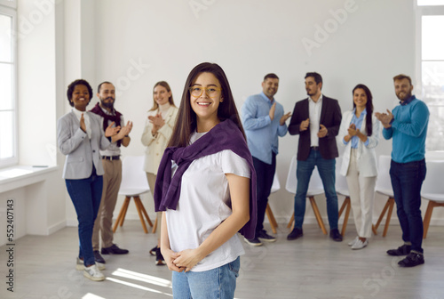Portrait of confident happy young woman in casual clothes against background of other people. Office worker or company manager working in business center posing against backdrop of applauding people.