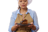 Selective focus on piece of homemade chocolate cake in hands of chef pastry, baker confectioner, wearing apron and hat blowing out a candle on birthday party, isolated on white background. Copy space