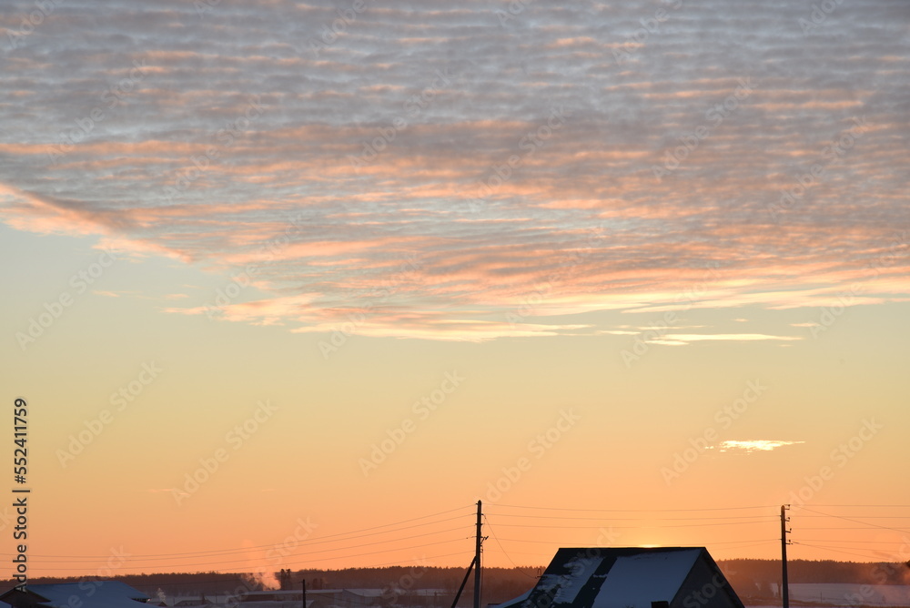 Sunset landscape in the countryside in winter. The setting sun behind the roof of the house in the evening. The disk of the sun and the evening sunset. Yellow pink beautiful sunset and sky.