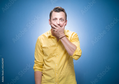 Oops. Ashamed young man with hand on mouth. Shame concept over blue background, dresses in yellow shirt