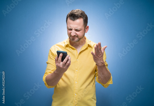 Nervous man holding mobile phone over blue background, dresses in yellow shirt. No Wi-fi concept. Bad connection, broken network low signal